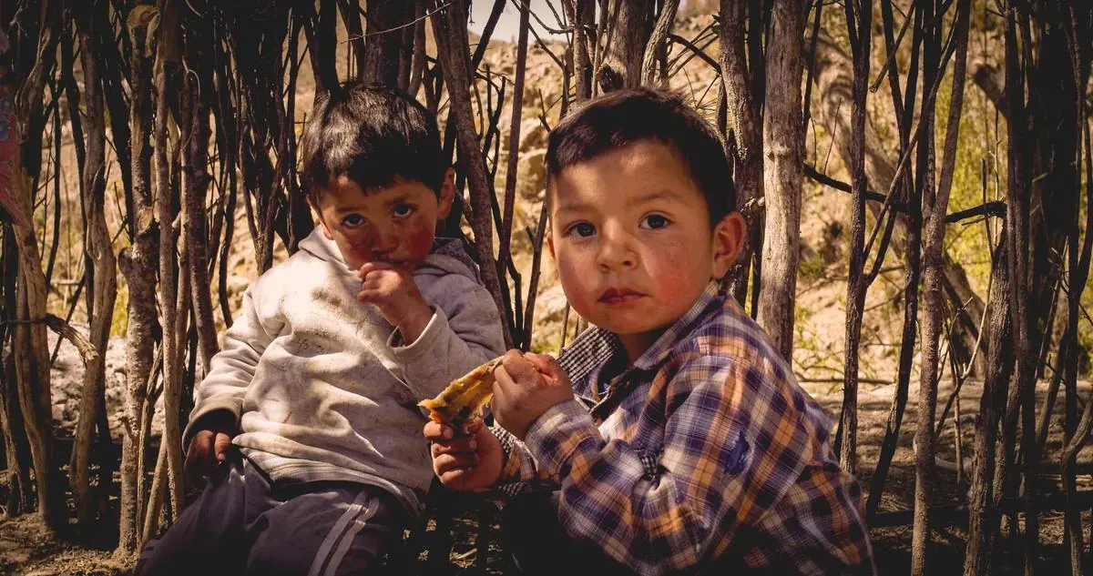 Dos chicos comiendo, sentados contra un cerco de ramas
